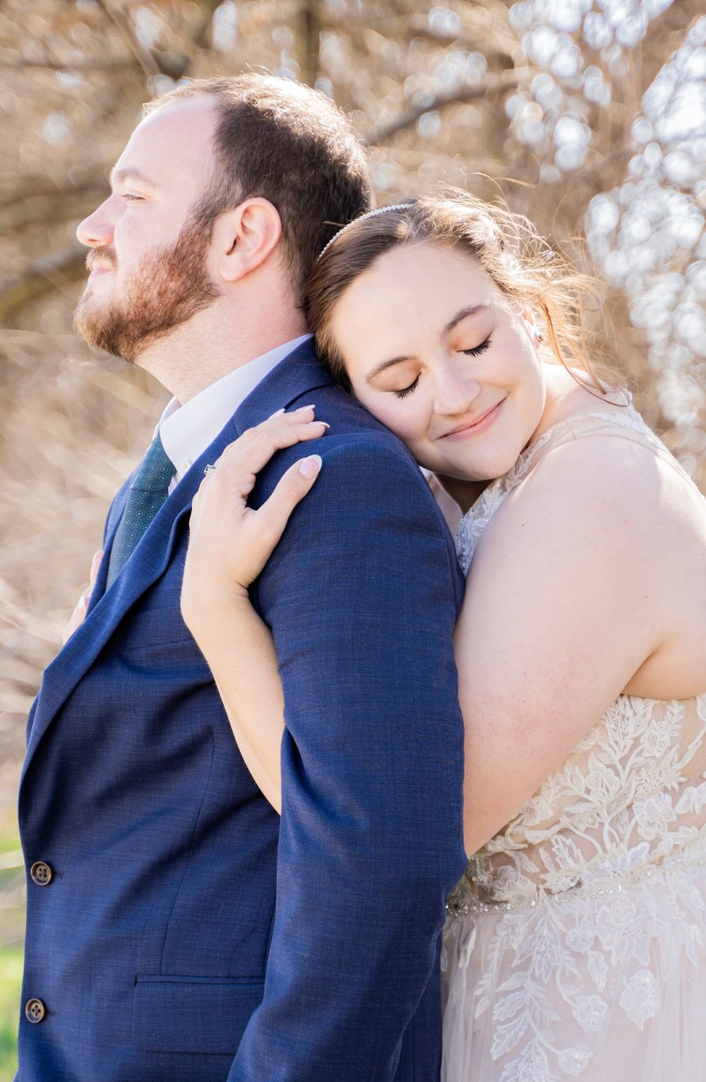 Bride in lace gown rests her head on grooms shoulder in a peaceful outdoor wedding portrait.