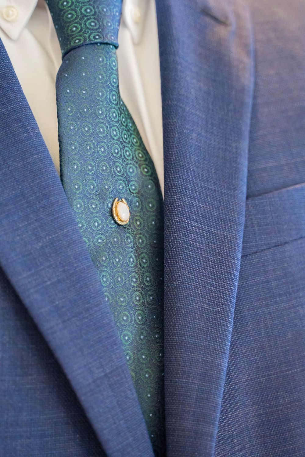Close-up detail of groom's navy blue suit with green patterned tie and gold tie pin accessory.
