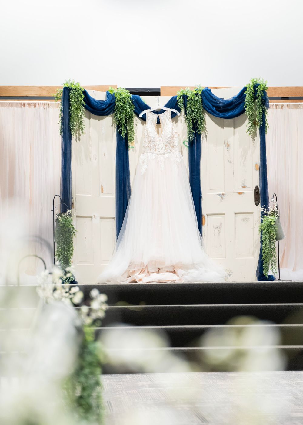 Lace wedding dress hanging in a decorated church with blue draping and greenery.