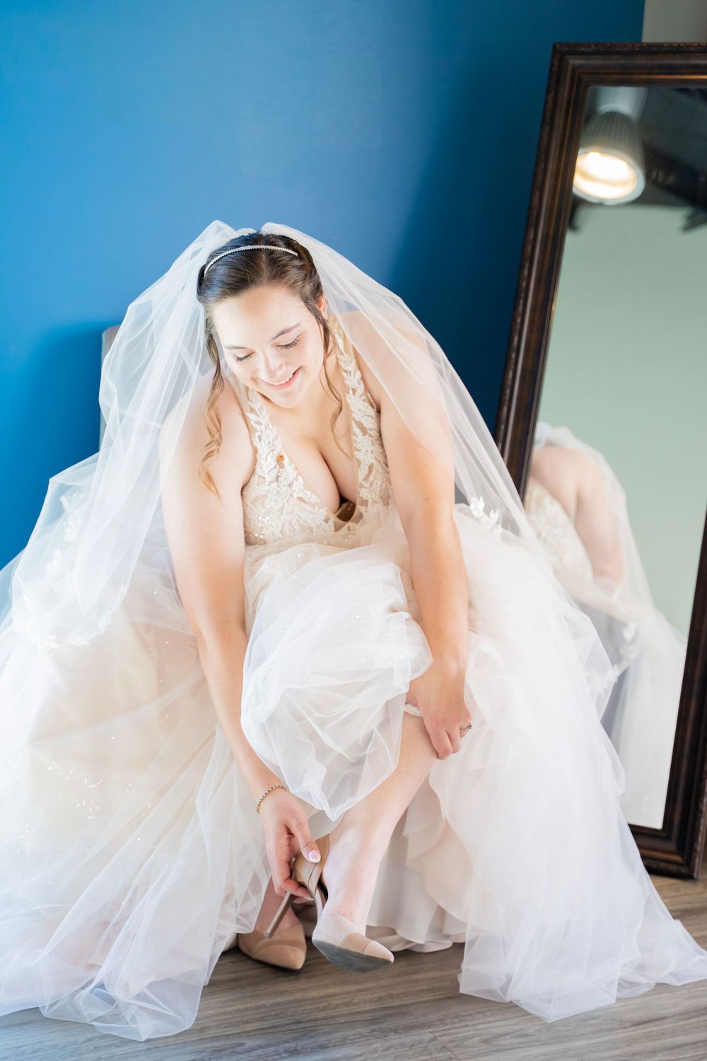 Smiling bride in white tulle gown and veil putting on nude heels near a mirror.