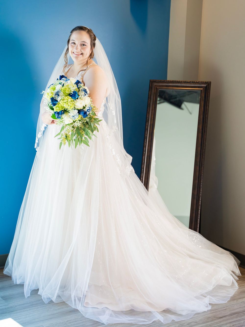 Bride in white ballgown and veil holding blue and green bouquet beside tall mirror.