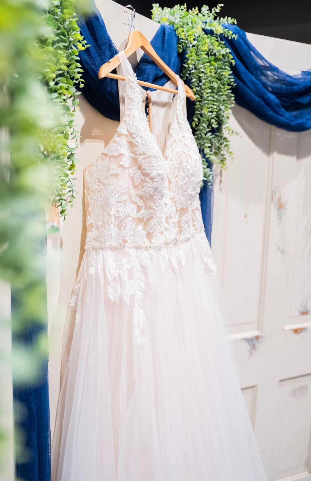 Lace and tulle wedding gown hanging on wooden hanger with blue floral backdrop.