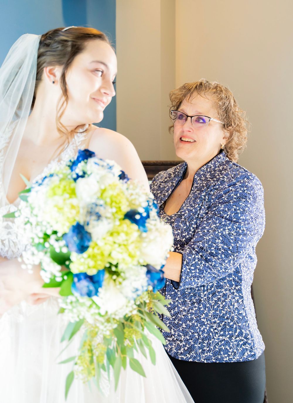 Bride in lace gown holding blue and white bouquet smiling with older woman in blue.