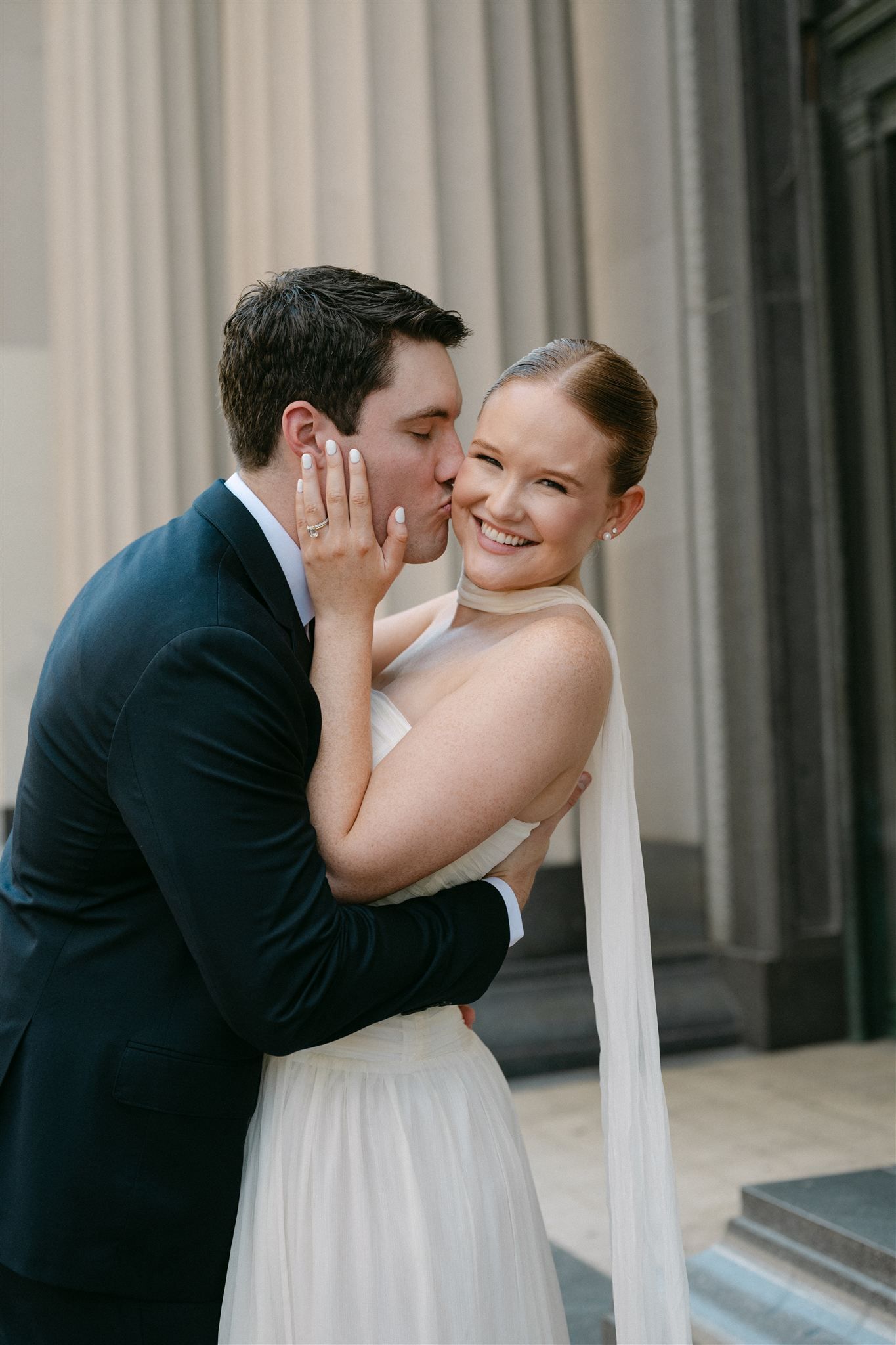 Newlywed couple hold each other and laughing while walking along city street with ornate columns behind them.