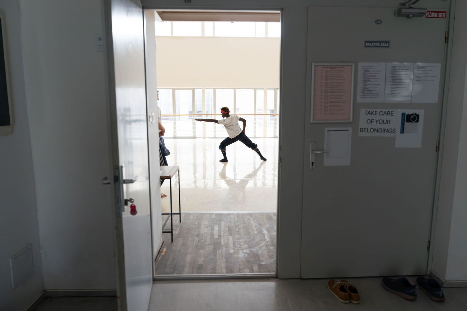 A dancer mid-lunge performs in a bright studio, viewed through an open doorway with notices on the wall.