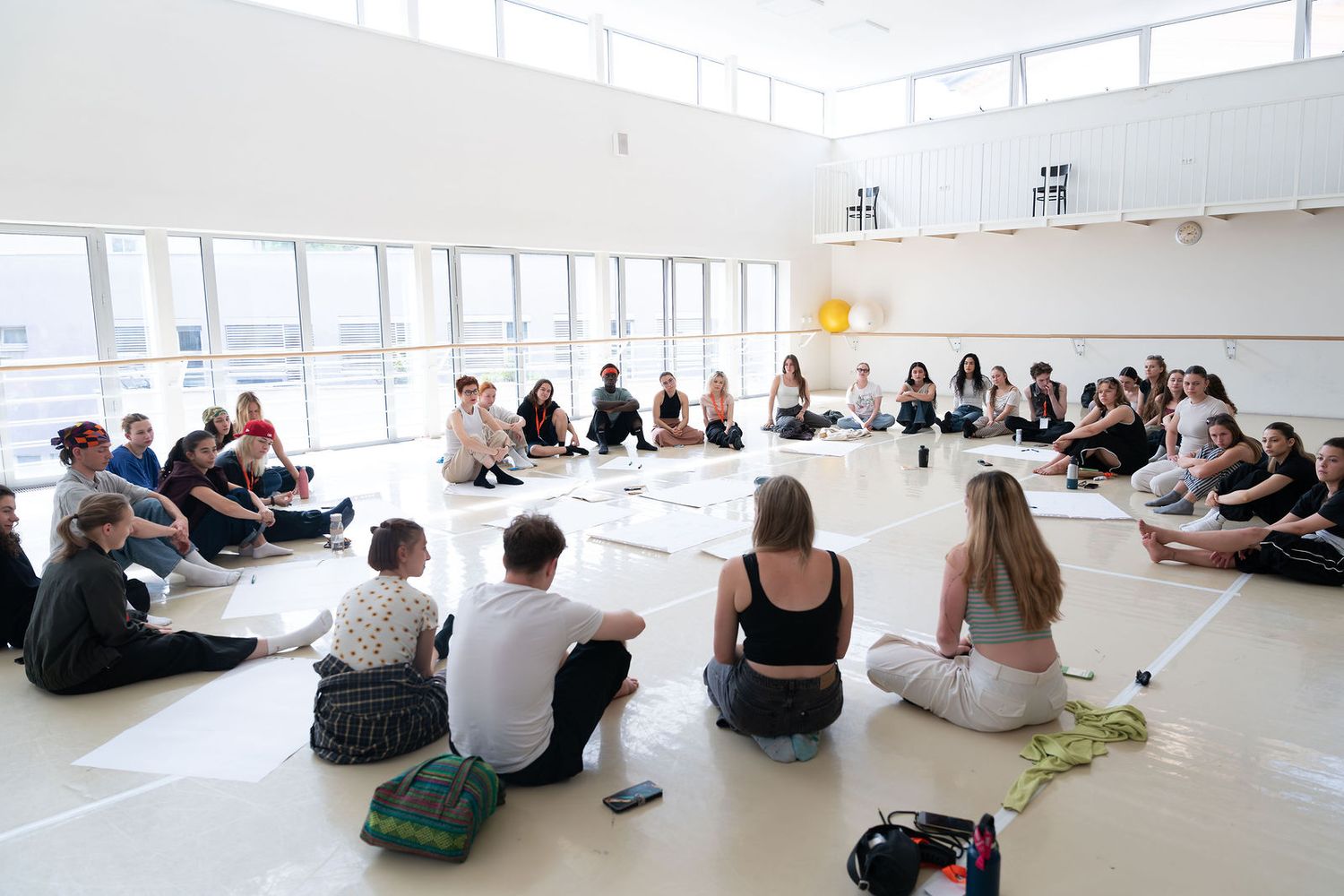Large group of people sitting in a circle on the floor of a bright, spacious dance studio during a workshop.