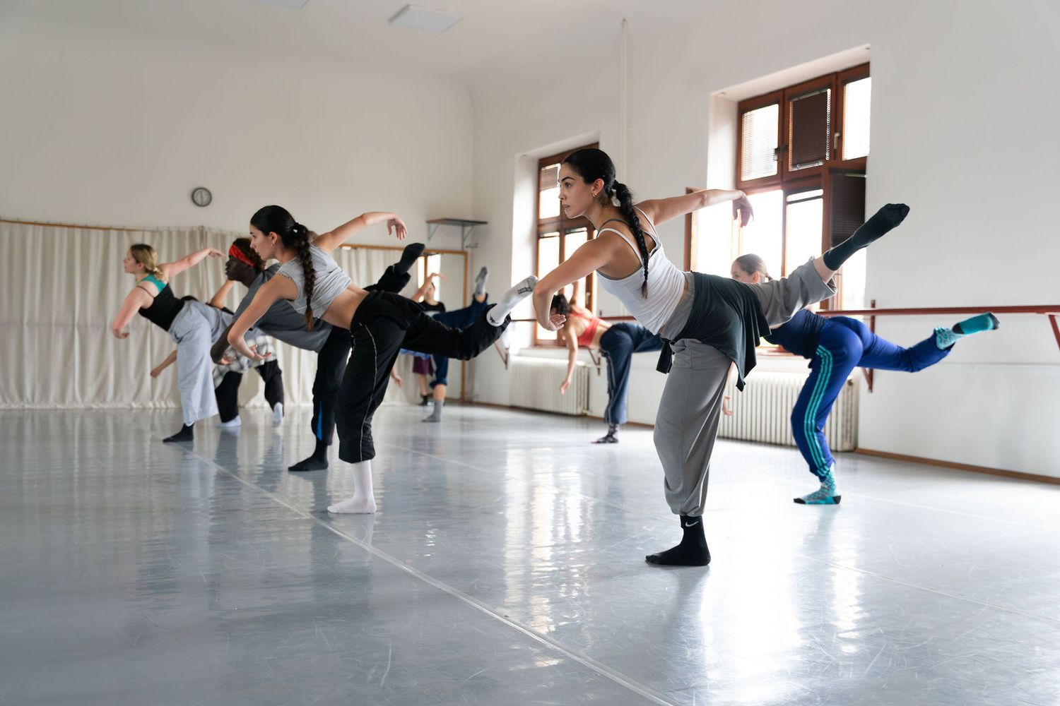 Dance students practice high kicks in a bright studio with ballet barres and large windows.