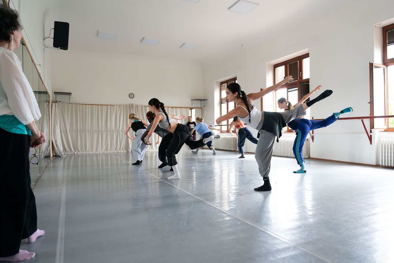 Dancers practice dynamic movements in a bright studio with mirrors, wooden floors, and large windows during a dance rehearsal.