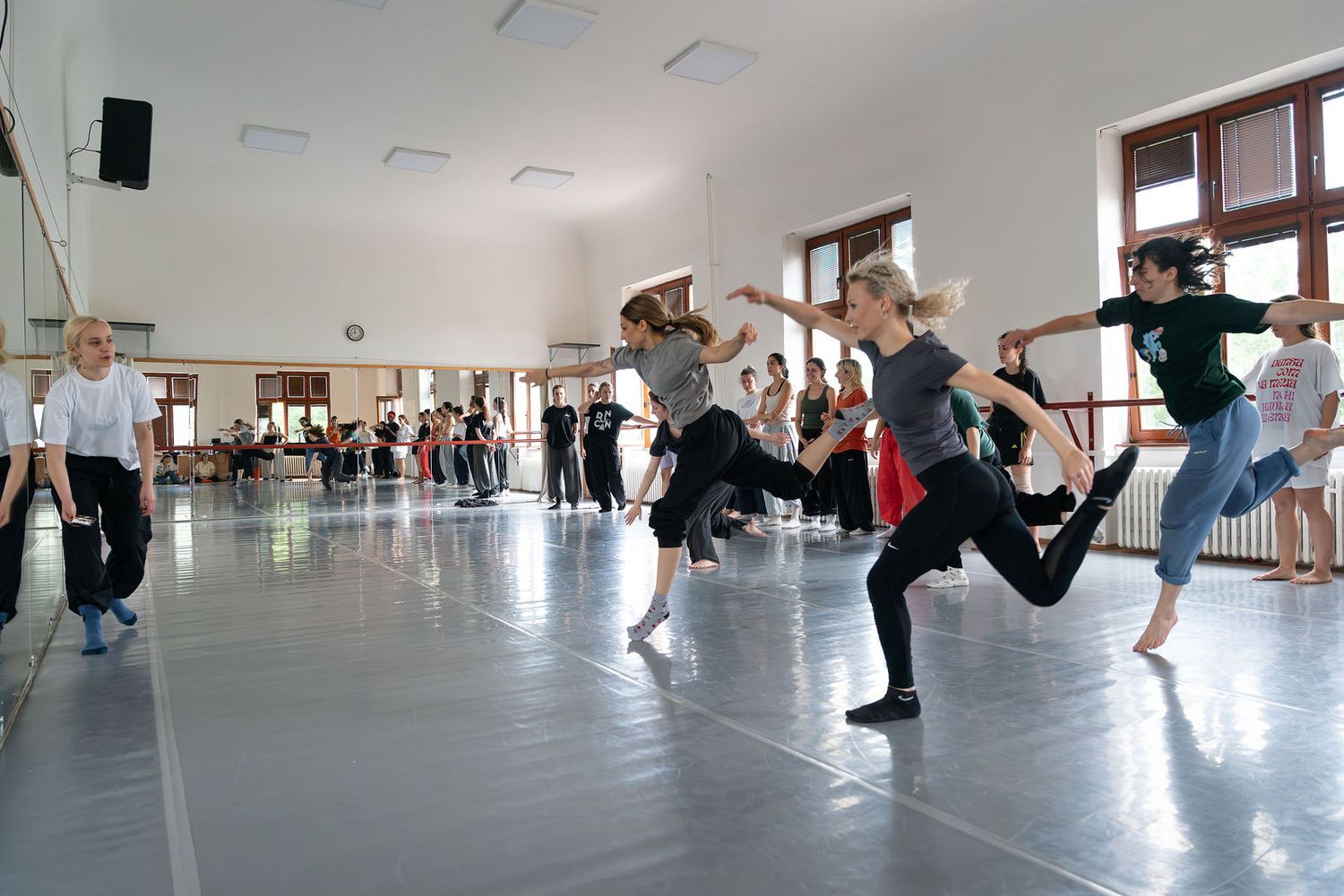 Dancers rehearse in a bright studio with mirrors, performing dynamic leaps and kicks during a dance class.