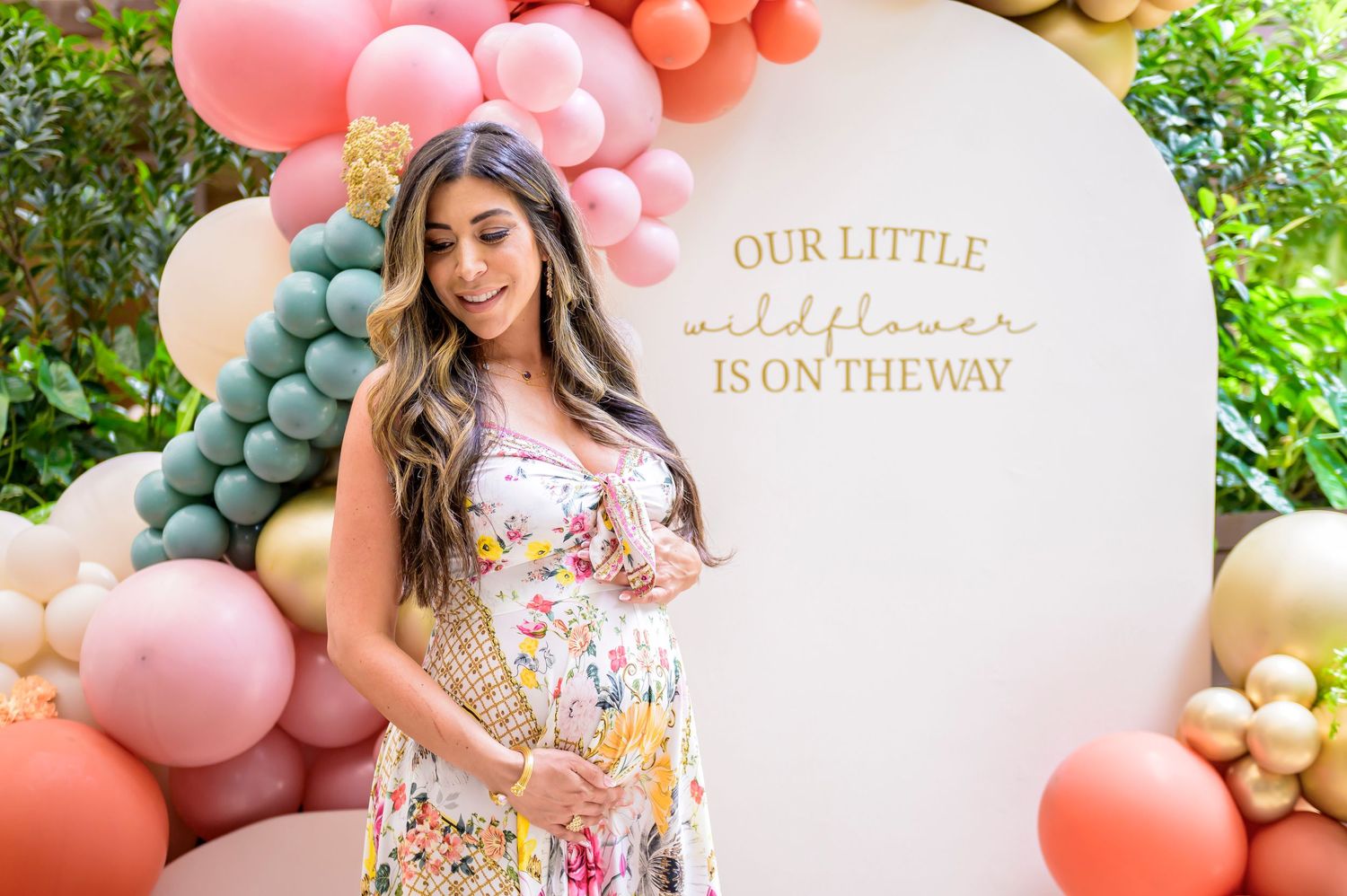 A maternity photoshoot setup with colorful balloon arch and text 'Our Little Is On The Way' against an outdoor garden backdrop.