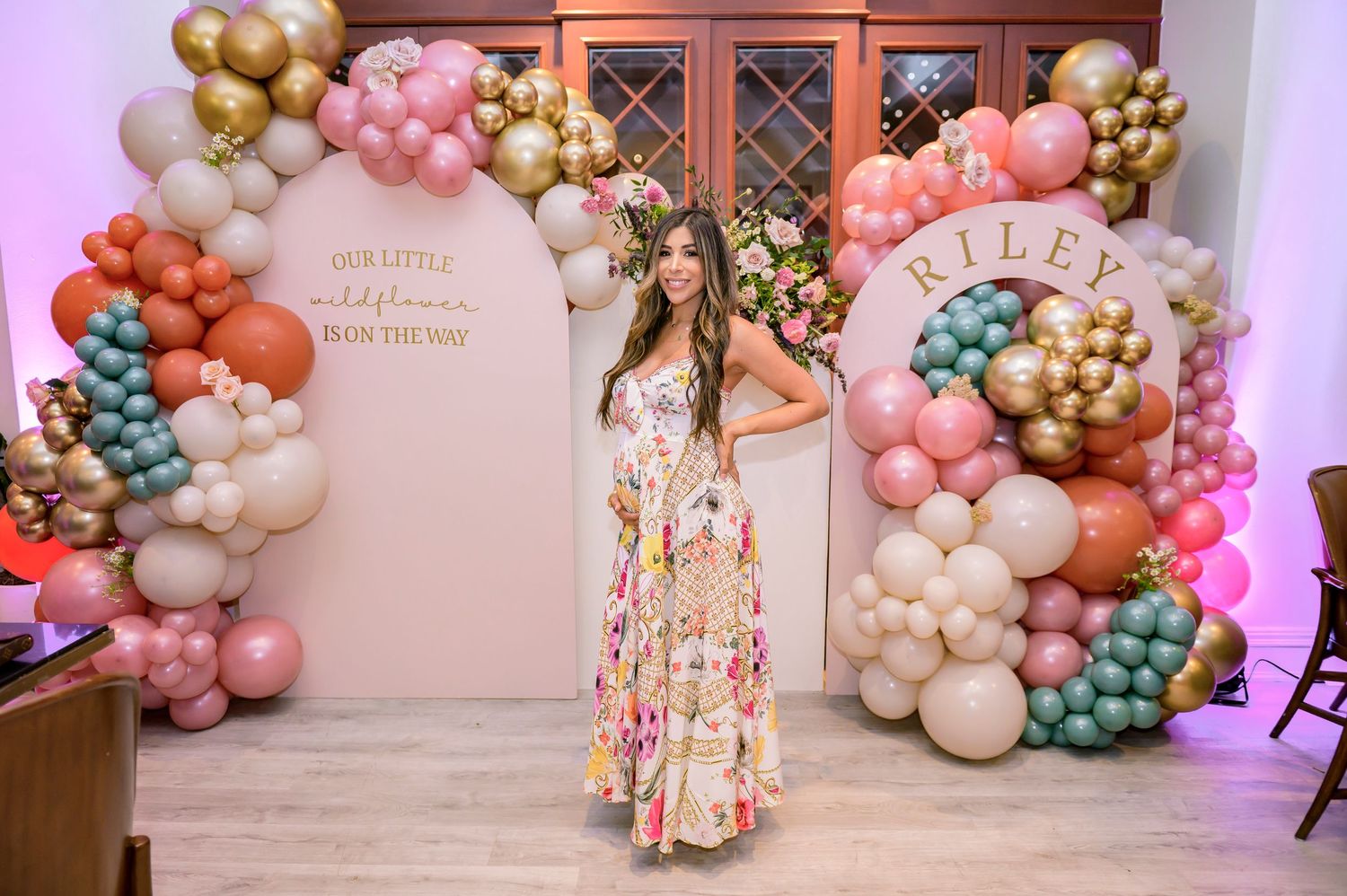 A mom-to-be in a floral dress posing at her baby shower with pink and gold balloon arches and decorative wall panels.
