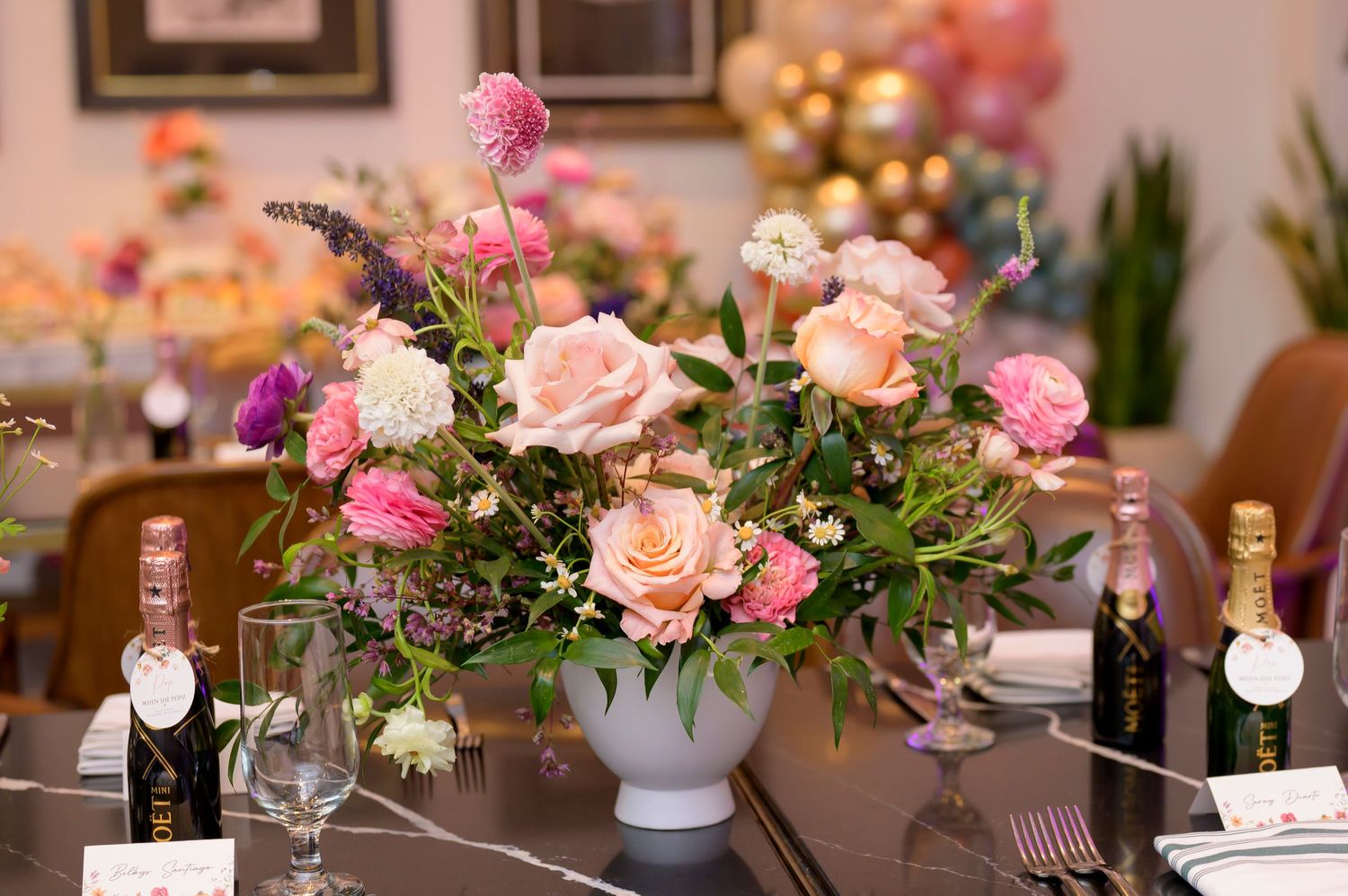 Pink and peach roses with purple flowers in a white vase centerpiece on elegant dining table with wine bottles.