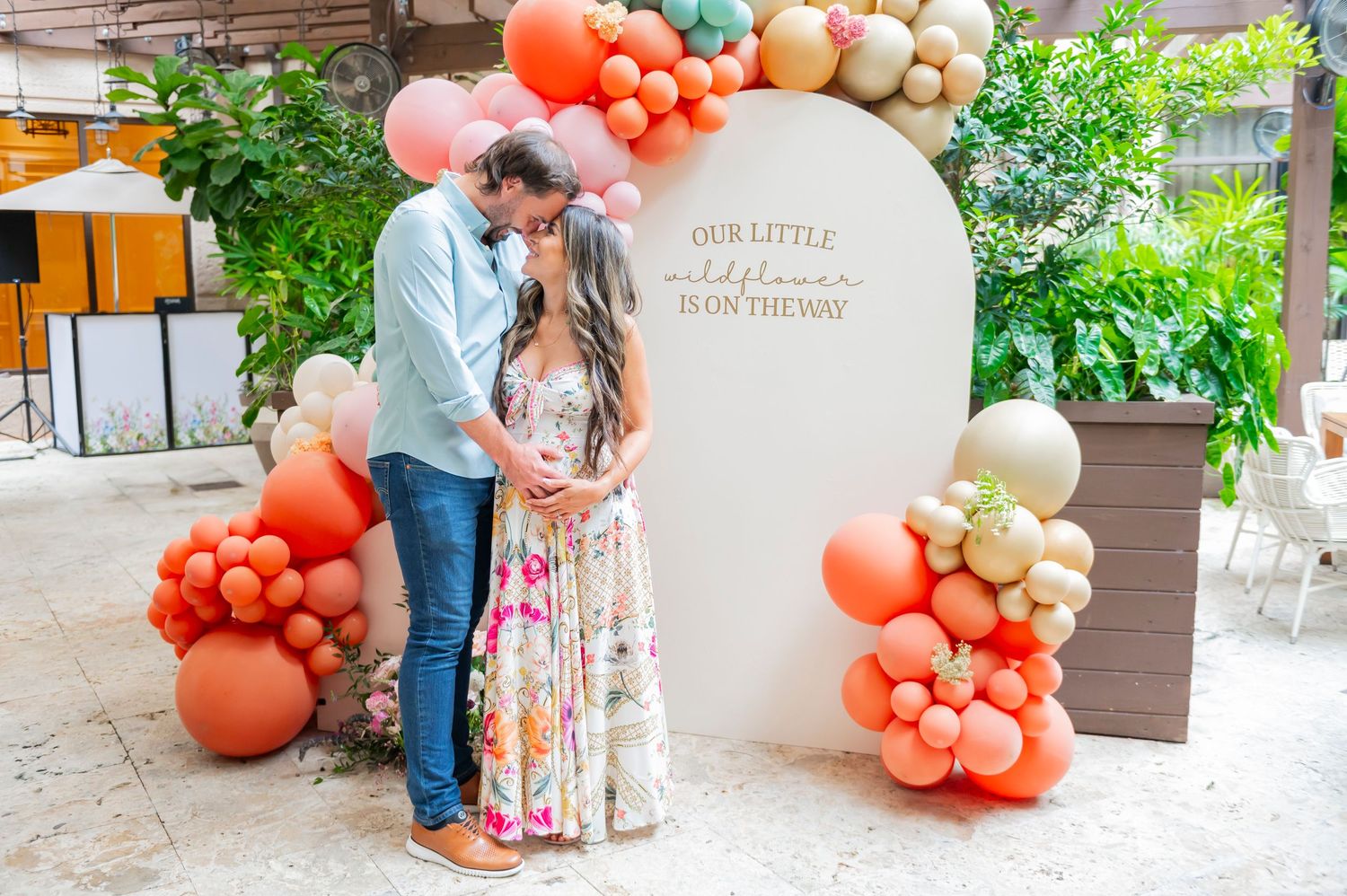 A romantic celebration setup with coral and beige balloon arrangements framing a circular backdrop in an outdoor garden setting.