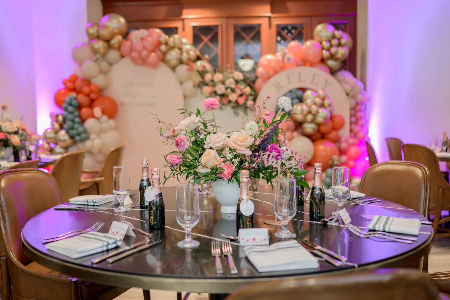Elegant table setting with floral centerpiece and purple lighting against a backdrop of balloon arch decorations.