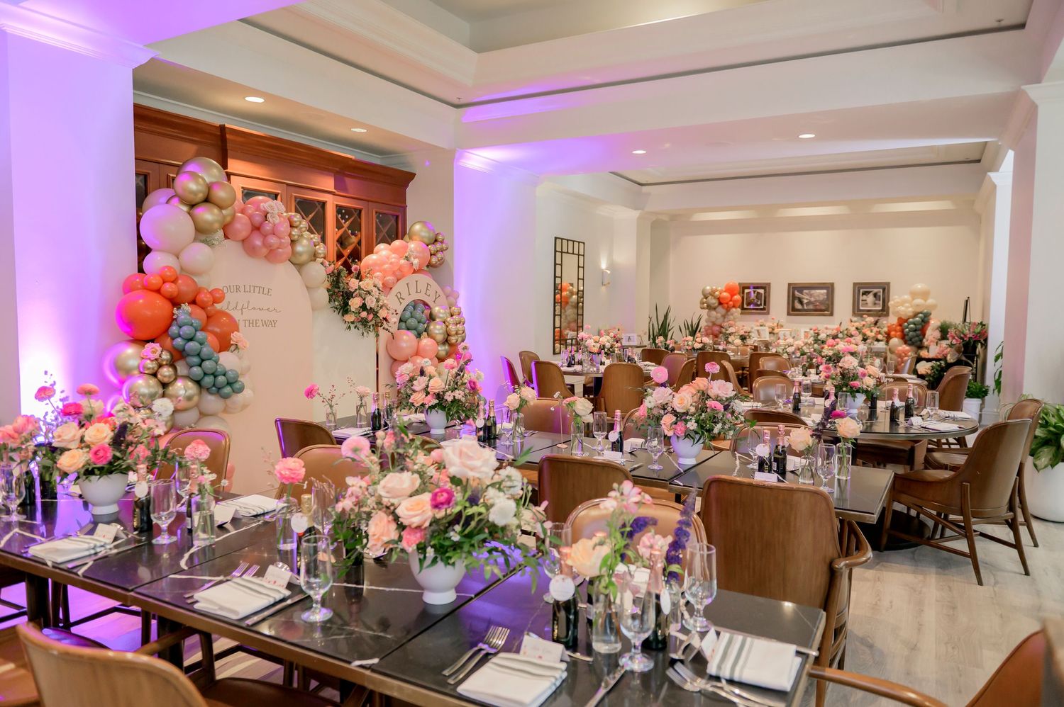 Elegant baby shower reception venue with floral arch, purple lighting, and table settings decorated with pink and white flowers.