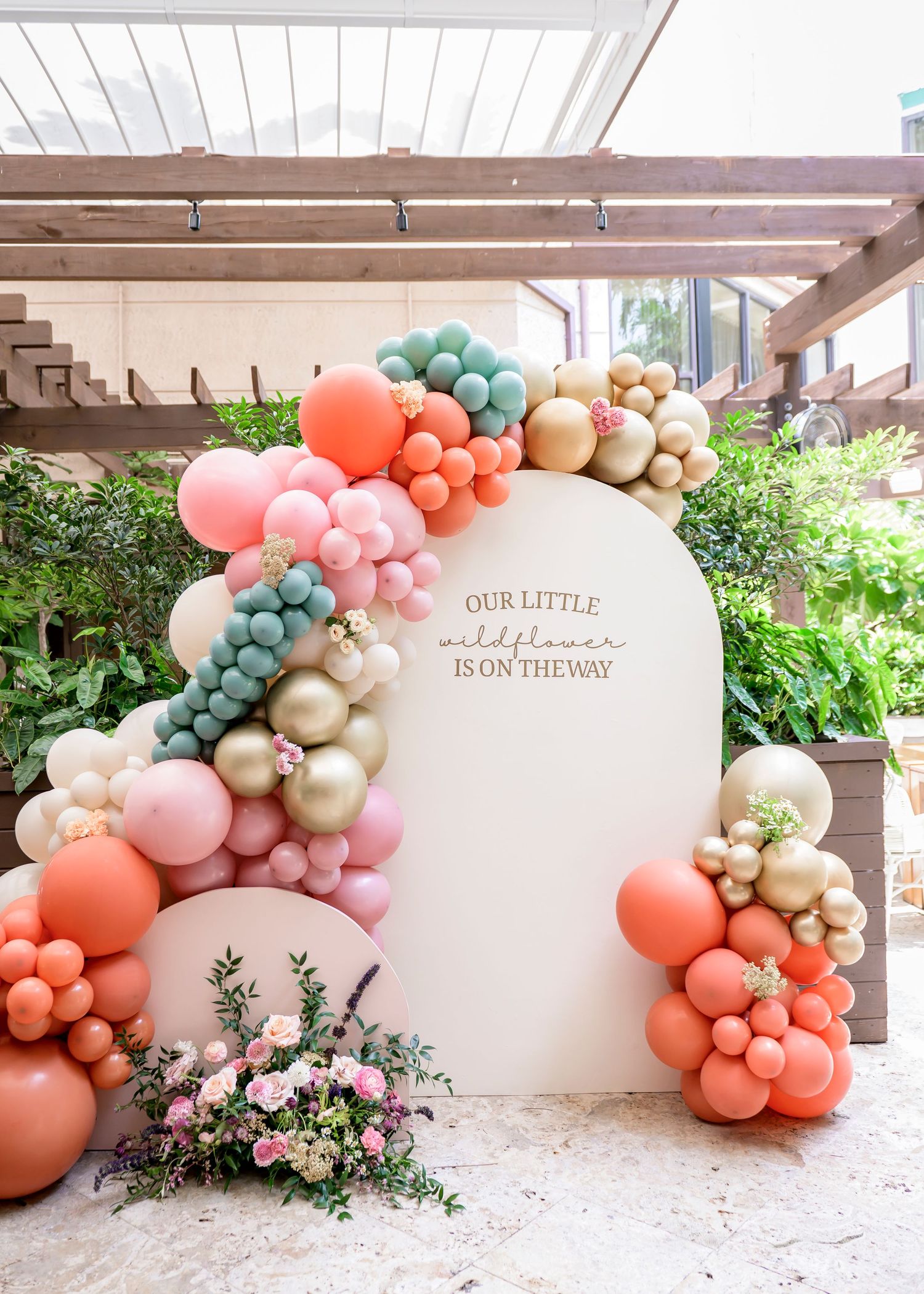 Elegant balloon arch with coral, sage, and gold balloons decorating a curved white backdrop with floral arrangements.