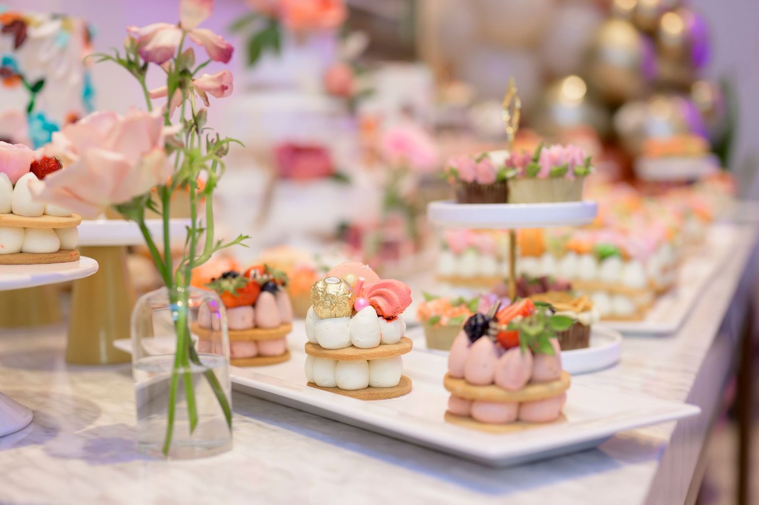 Elegant dessert table with pink macarons, floral arrangements, and delicate pastries displayed on white serving plates.