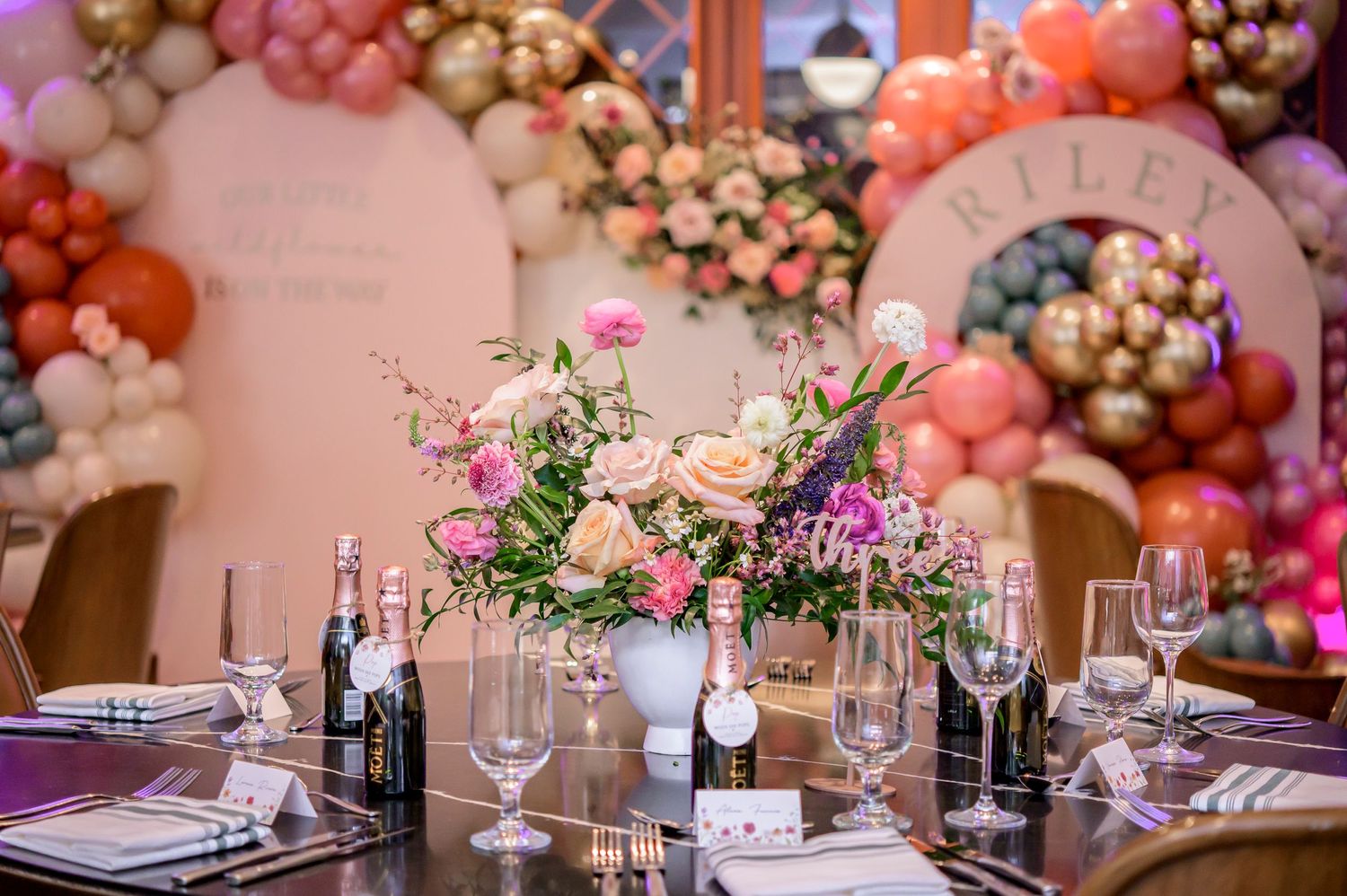 Elegant table setting with pink floral centerpiece and balloon arch backdrop at an upscale event venue.