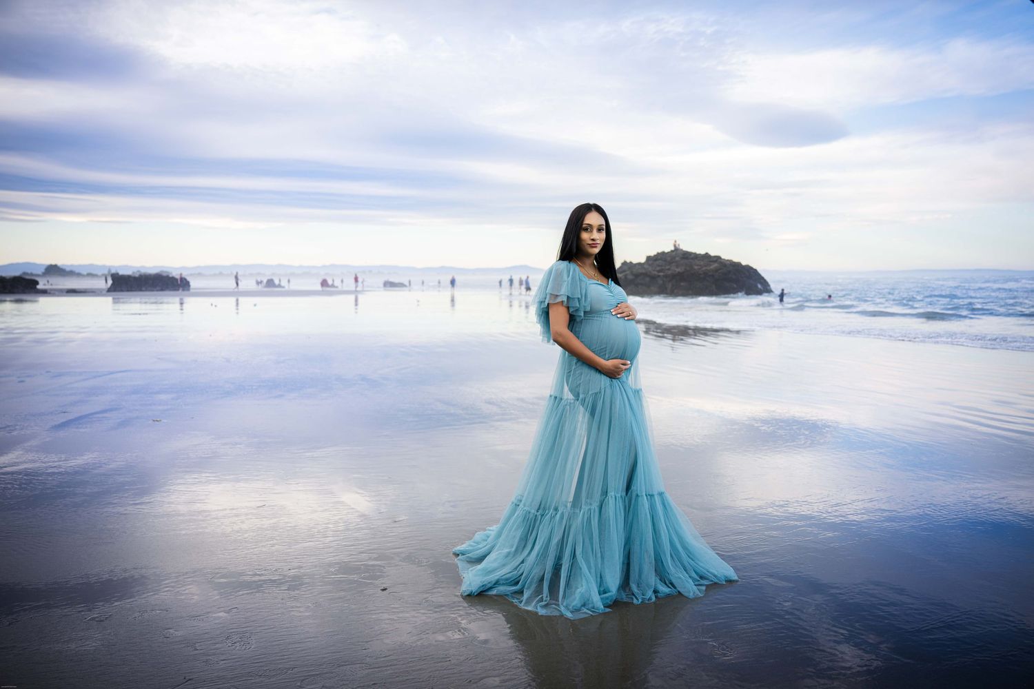 A flowing turquoise maternity gown creates an ethereal scene against the reflective sand and ocean waves at dusk.