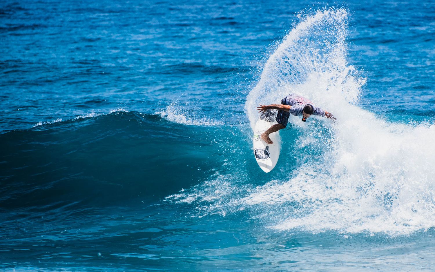 A surfer performs a powerful top turn maneuver on a blue ocean wave, creating a dramatic spray of white water.