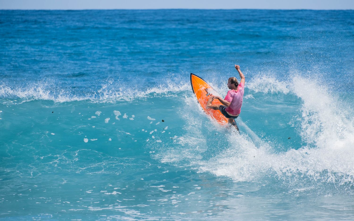 Surfer on an orange board performing a dynamic aerial maneuver on a turquoise wave.