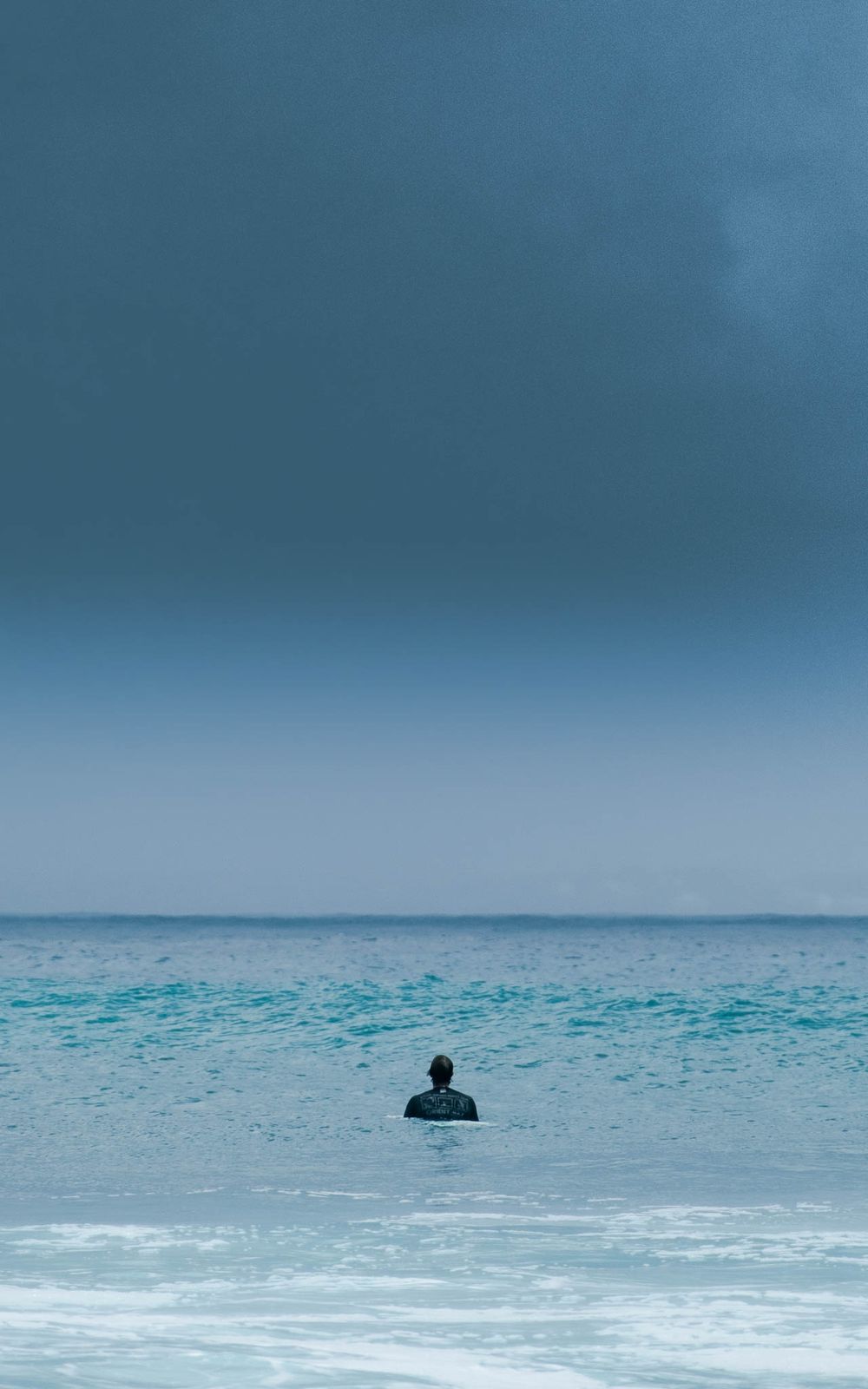 Lone surfer sits in calm ocean waters under moody grey skies.