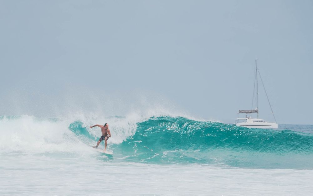 Surfer rides a turquoise wave with a sailboat visible in the background.