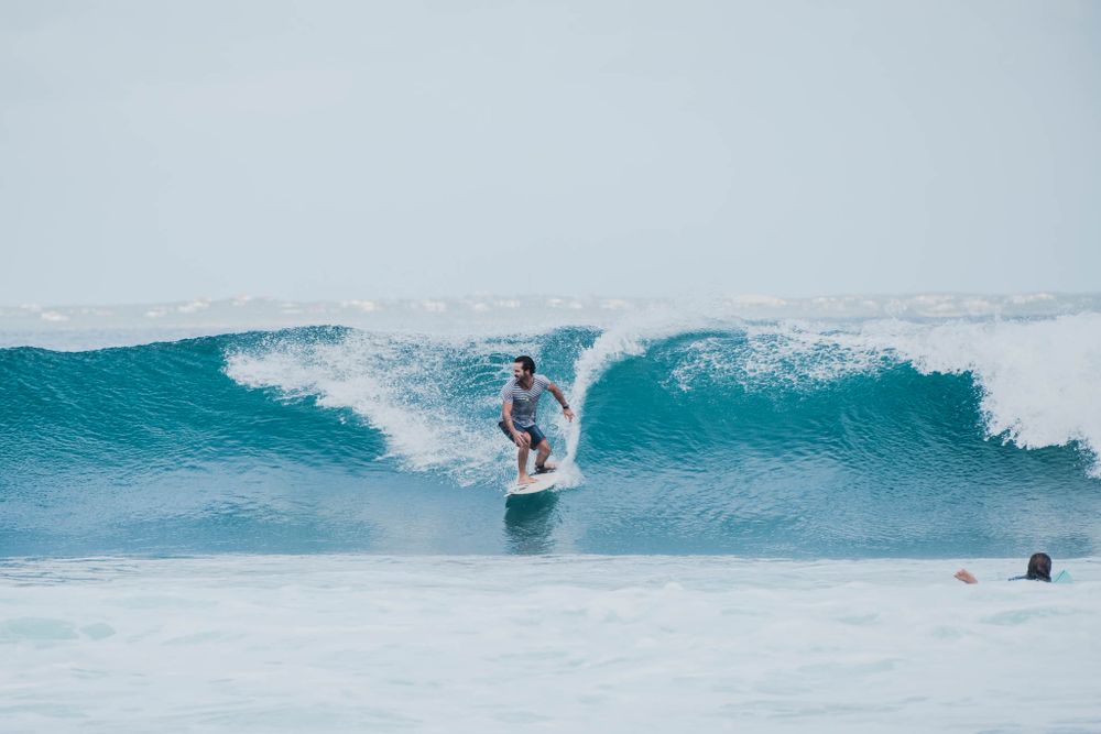 Surfer rides the face of a perfect turquoise wave under an overcast sky.