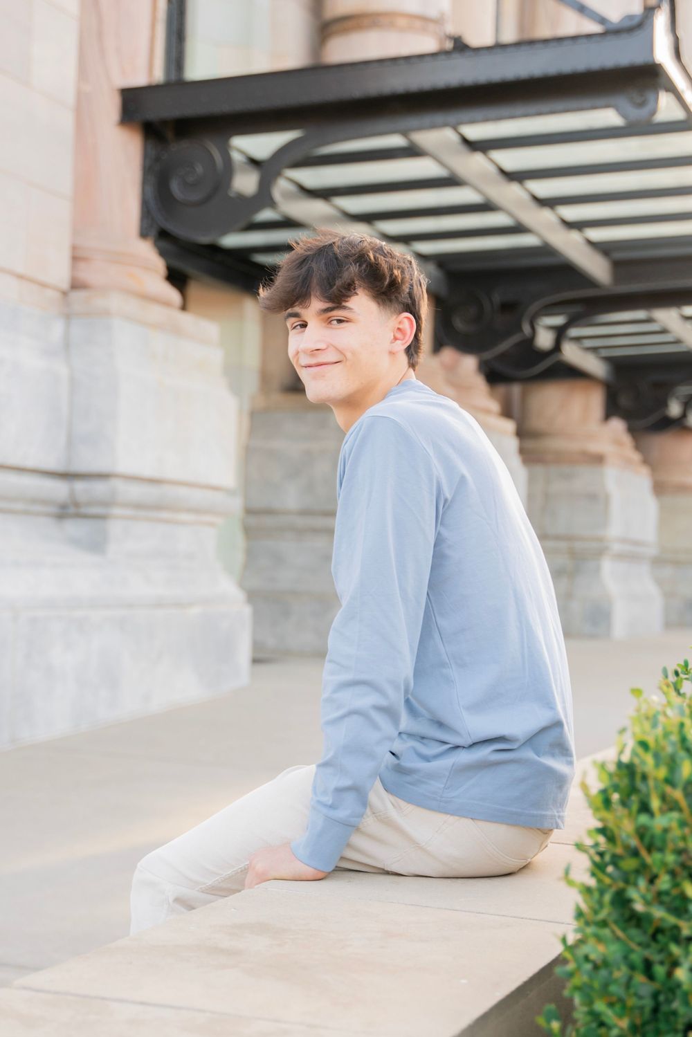 Smiling teen boy in blue long-sleeve shirt sitting on stone ledge outside historic building.