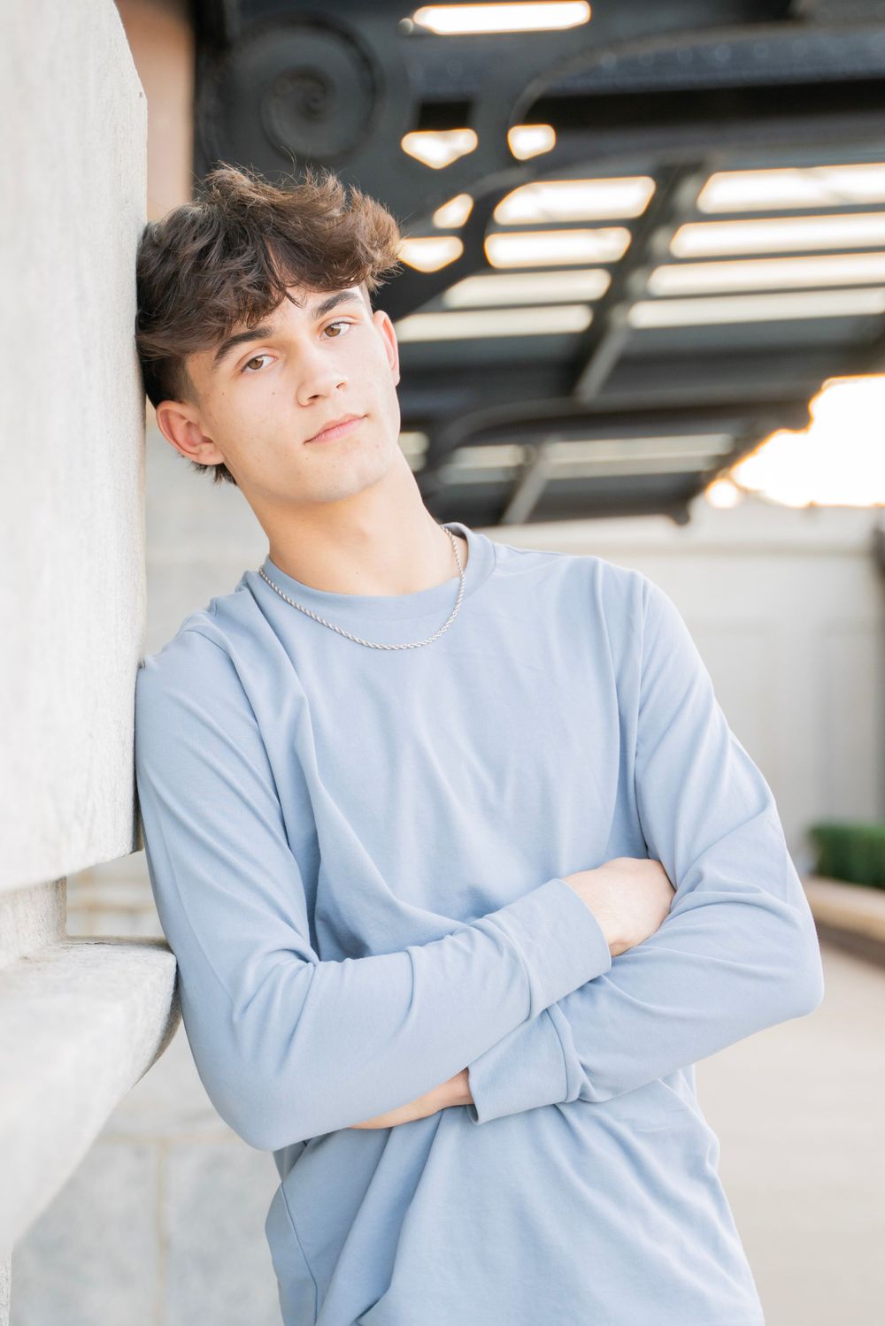 Confident teen boy in blue long-sleeve shirt leaning against column with arms crossed.