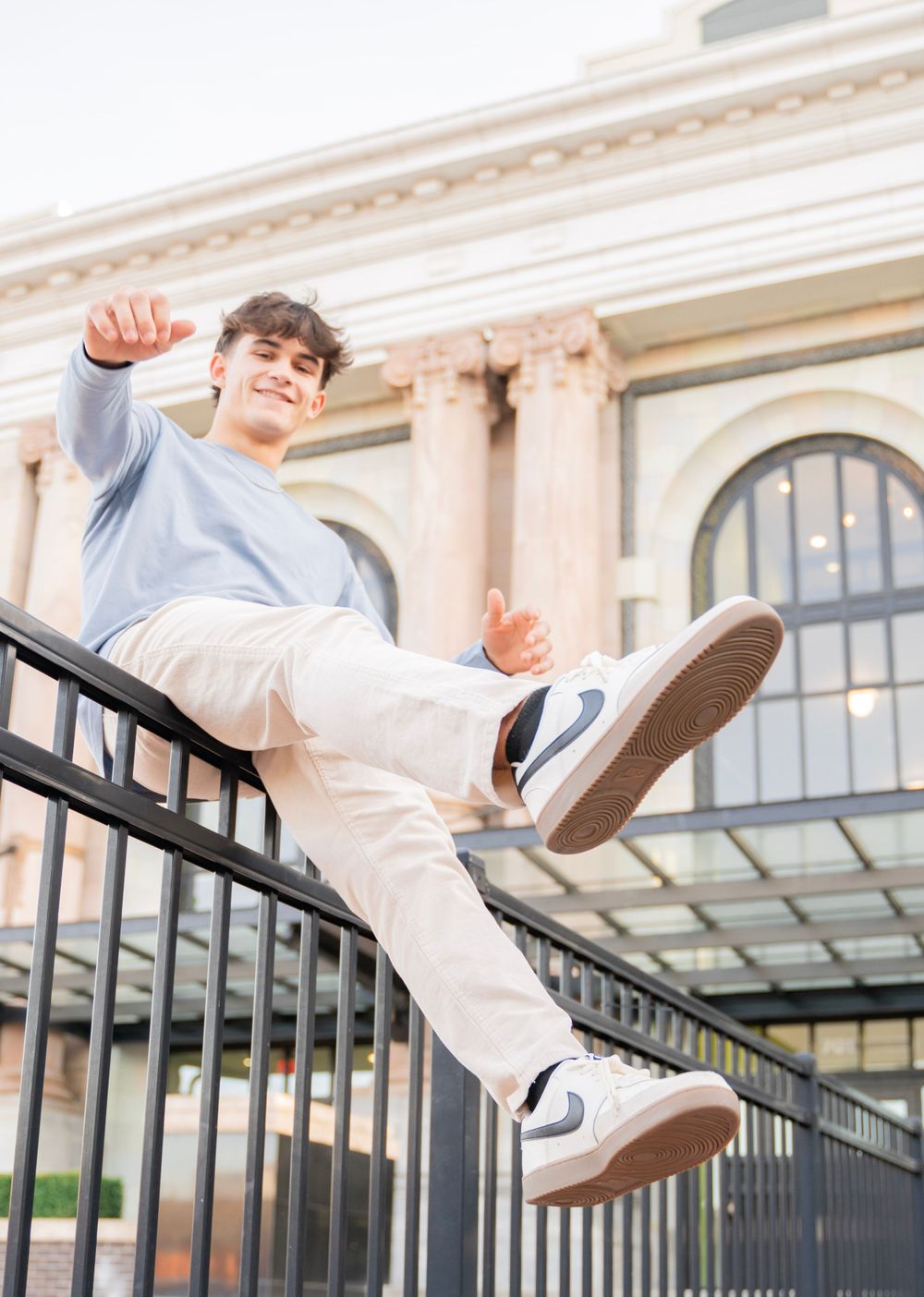 Young man in blue shirt and white Nikes balancing on black railing outside classic building.