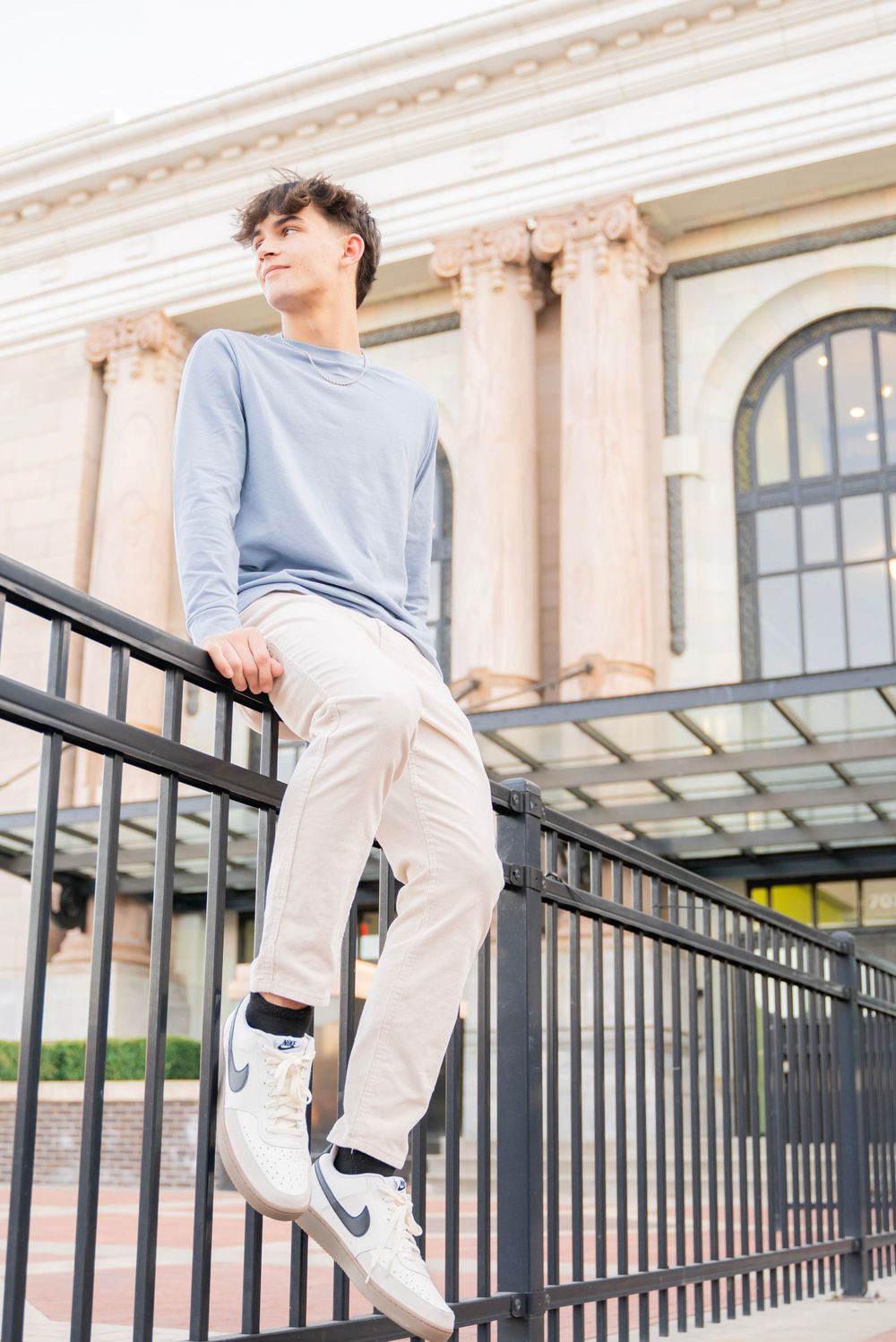 Teen boy in light blue long sleeve and cream pants leaning on black iron railing outdoors.