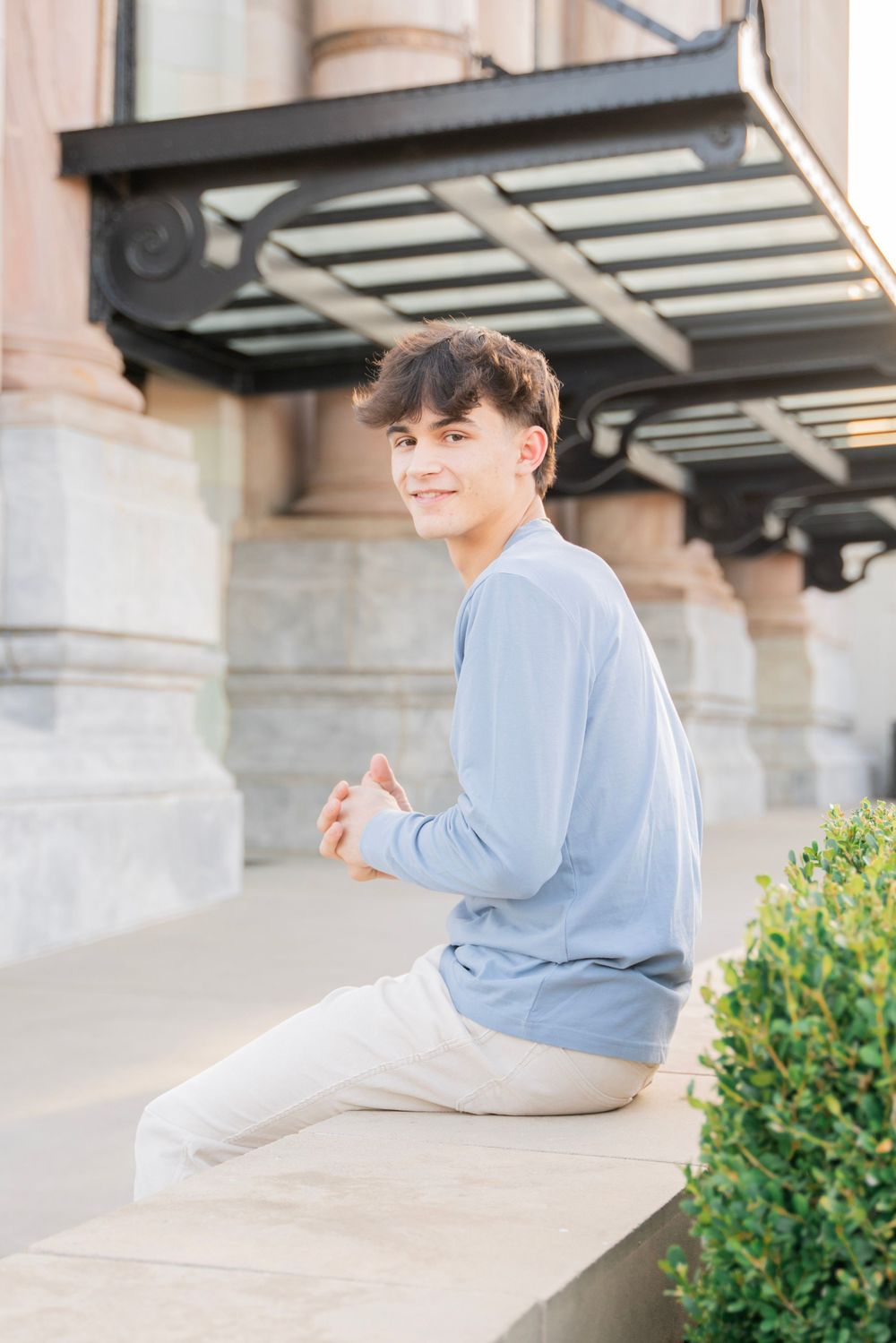 Young man in light blue sweater and khakis sitting on stone ledge near classic architecture.
