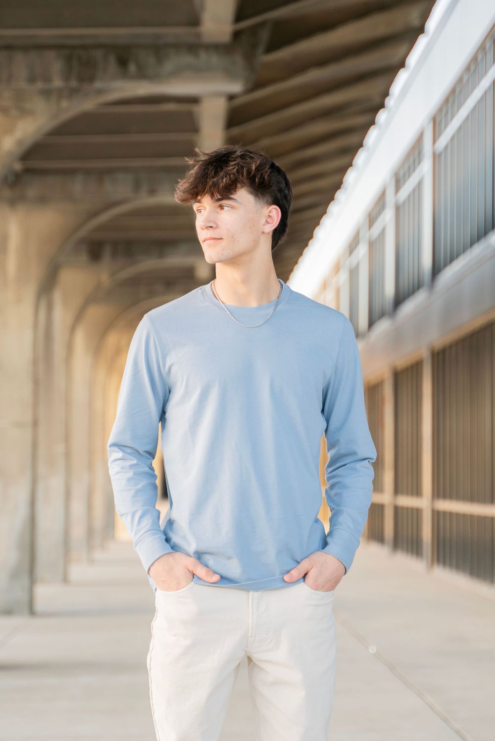 Young man in light blue long sleeve shirt standing in urban corridor with hands in pockets.