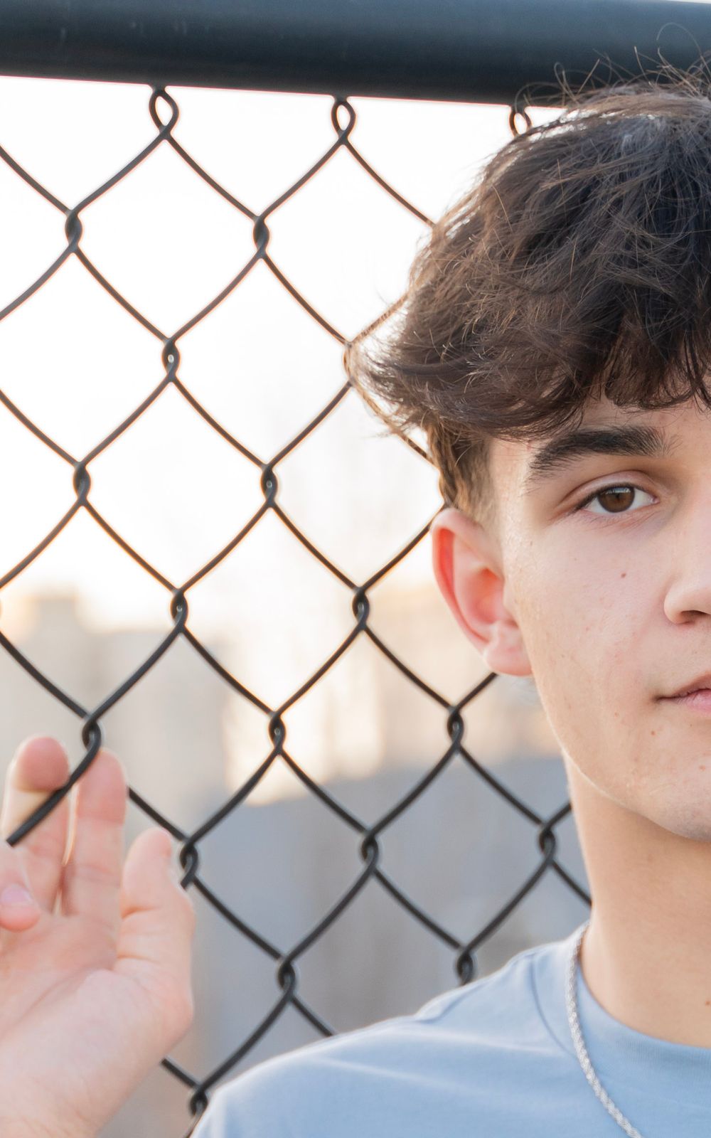 Close-up portrait of teenage boy with dark hair looking through chain-link fence outdoors.