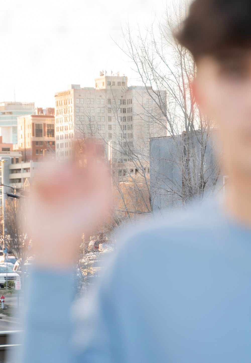 Blurred back view of person in blue shirt with urban cityscape and bare trees in background.