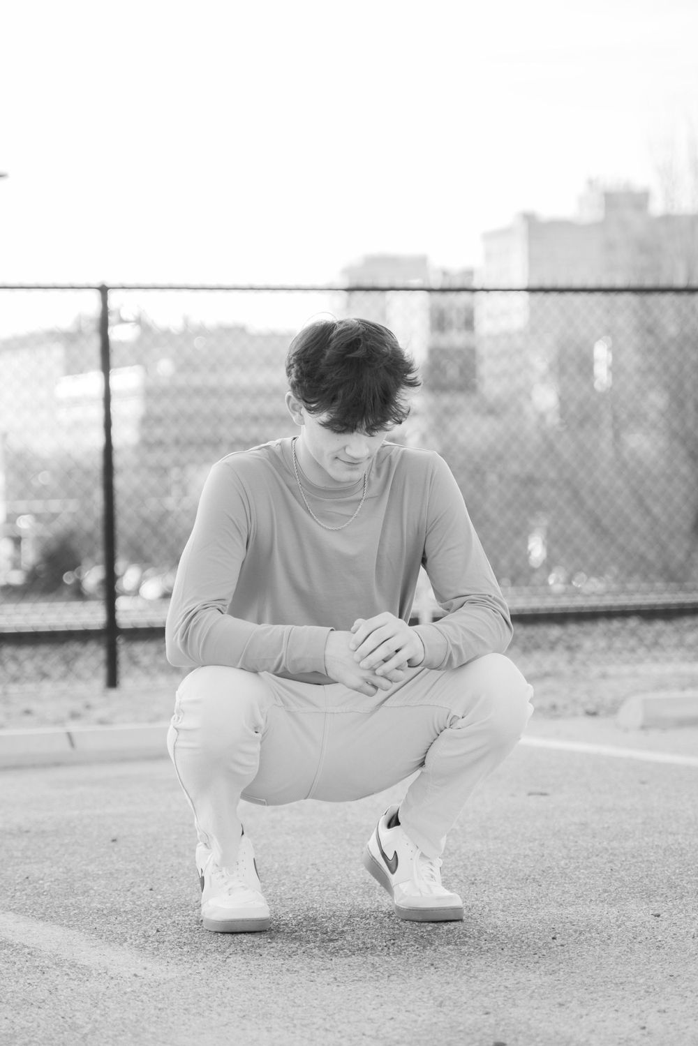 Black and white photo of young man crouching on rooftop with chain-link fence behind him.