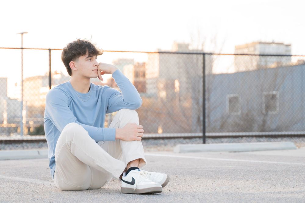 Young man in blue sweater and beige pants sitting on rooftop parking lot at golden hour.