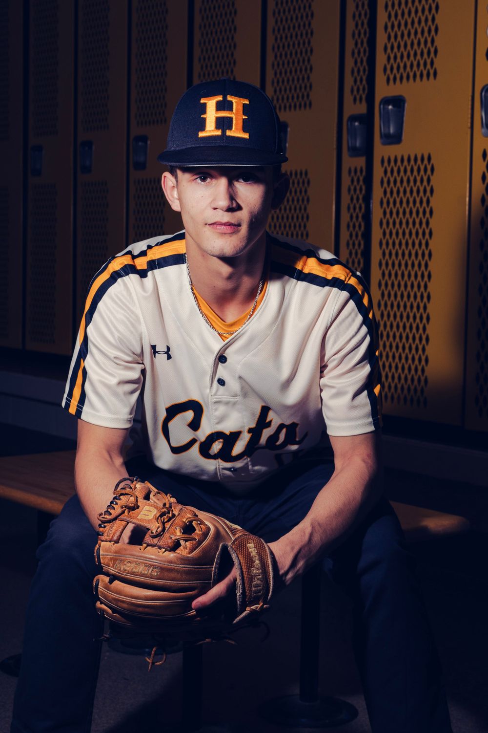 Teen baseball player in white Cats uniform holding glove sitting in school locker room.