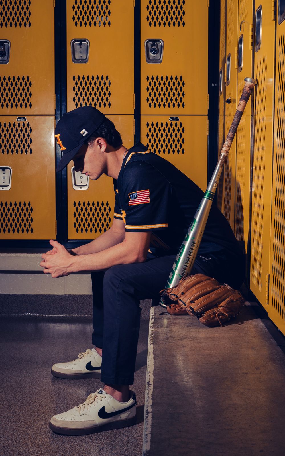 Teen baseball player in navy uniform sitting pensively in yellow school locker room.