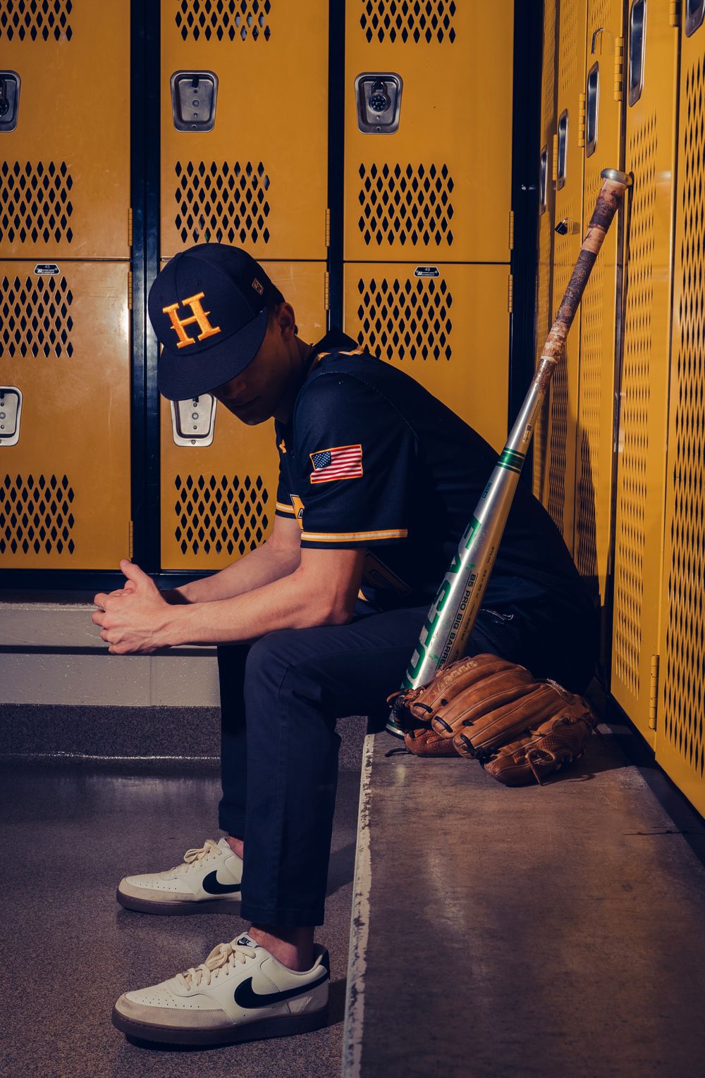 Baseball player in navy Cats uniform sitting in yellow locker room with bat and glove.