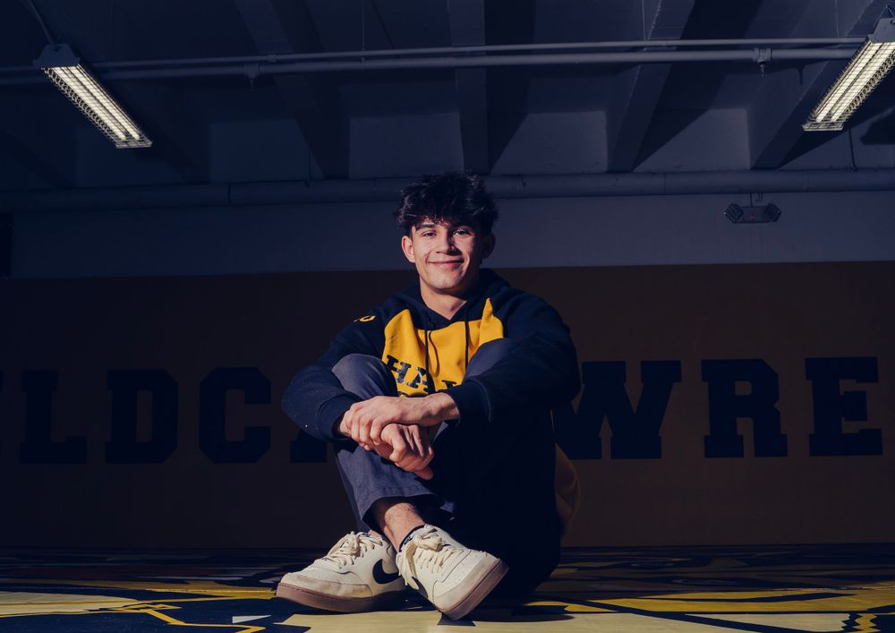 Teen boy in yellow and navy jacket sitting on wrestling gym floor with school banner behind.