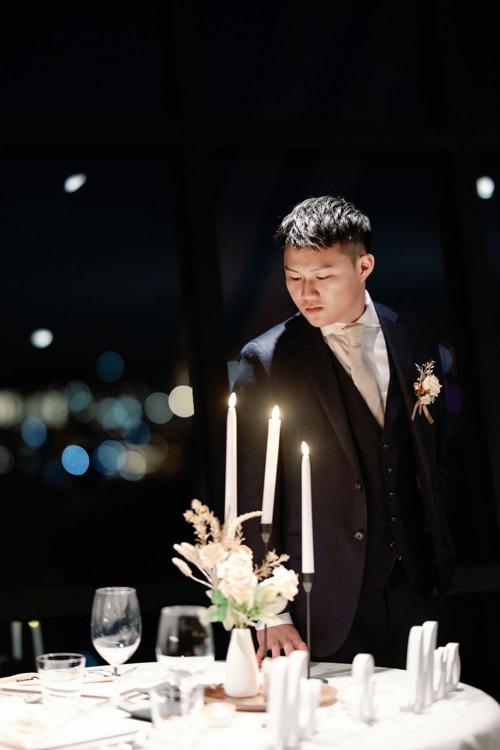 Groom in navy suit gazes at candlelit reception table with floral centerpiece at night wedding.