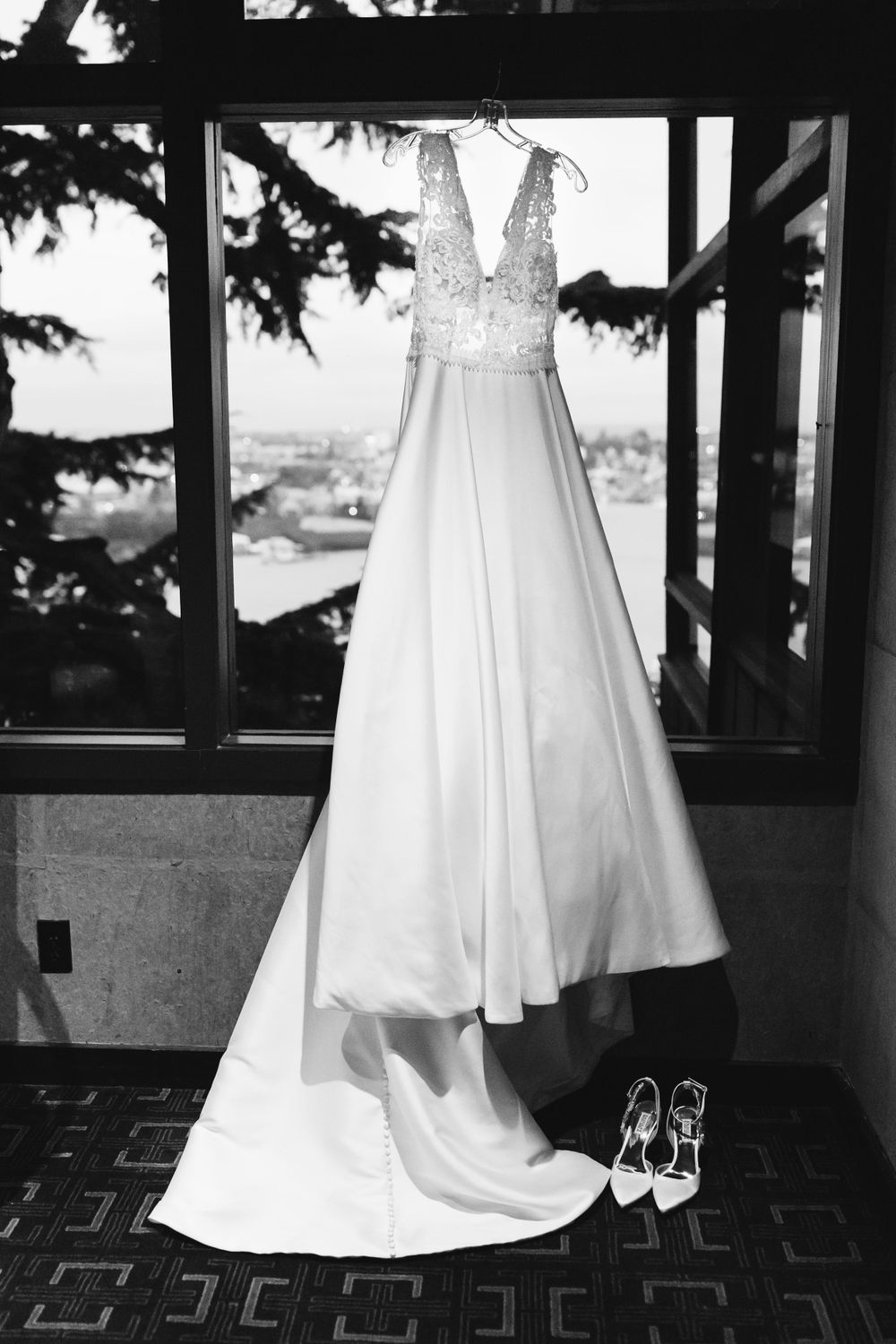 Black and white photo of an elegant wedding gown with lace bodice hanging by a window with bridal heels below.