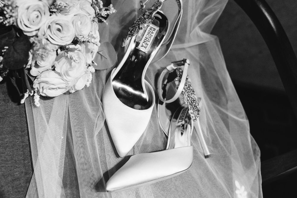 Black and white photo of elegant bridal heels beside a white rose bouquet and wedding veil.