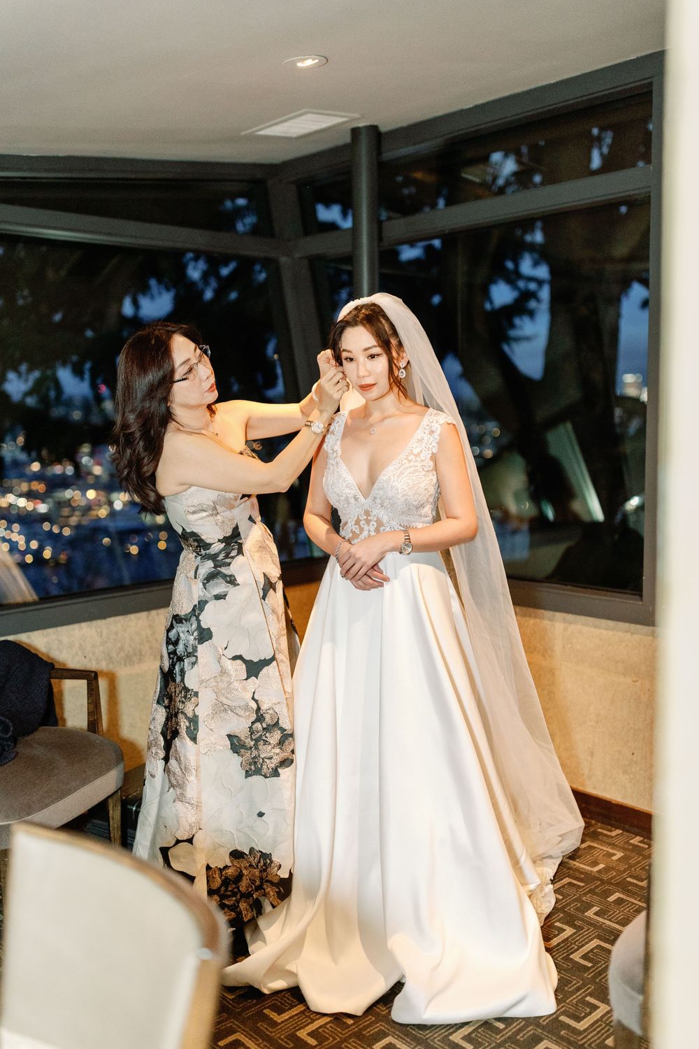 Mother helping bride with veil and accessories in a window-lit bridal suite before the wedding ceremony.