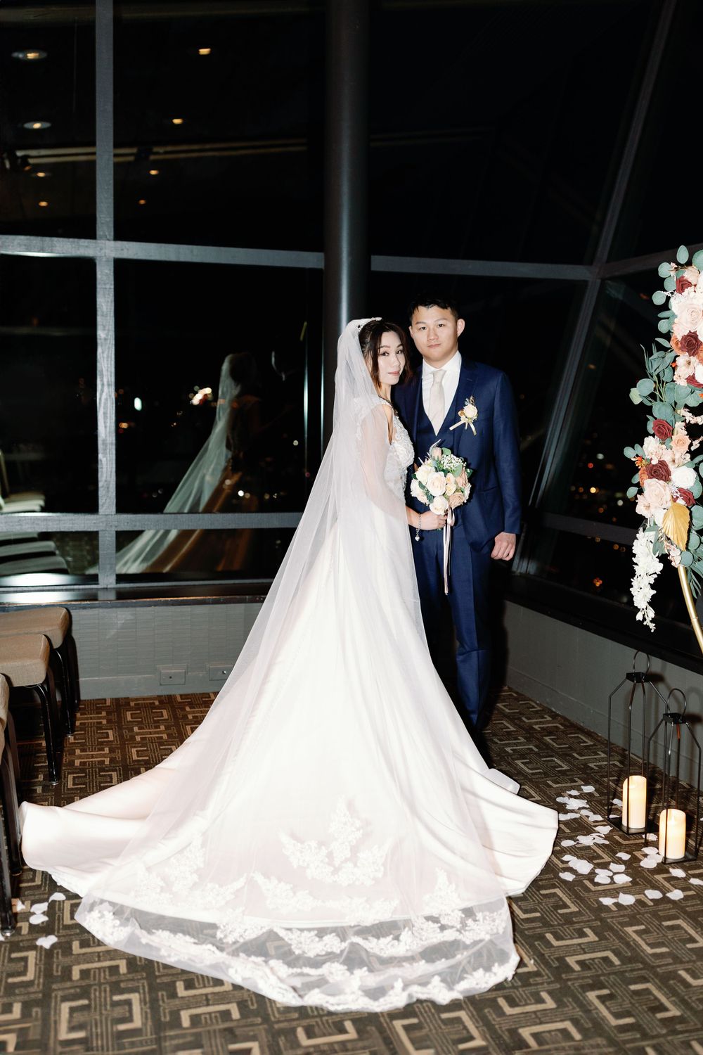 Asian bride and groom posing elegantly with long cathedral veil and floral arch at night.
