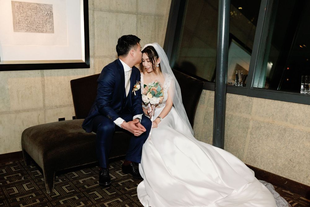 Bride and groom sharing an intimate moment seated together holding a bridal bouquet indoors.