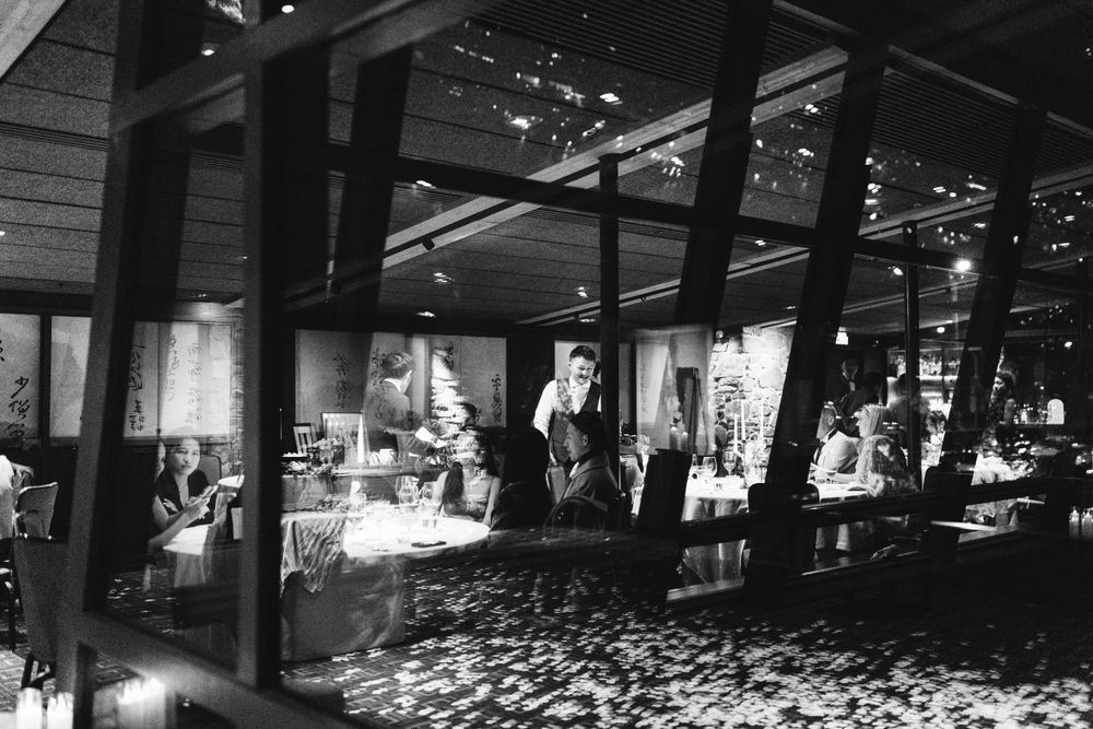 Black and white photo of an elegant restaurant interior with diners and a waiter serving guests in a dimly lit setting.