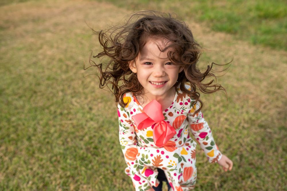 Young child in floral dress twirls joyfully on grassy lawn during outdoor photoshoot.